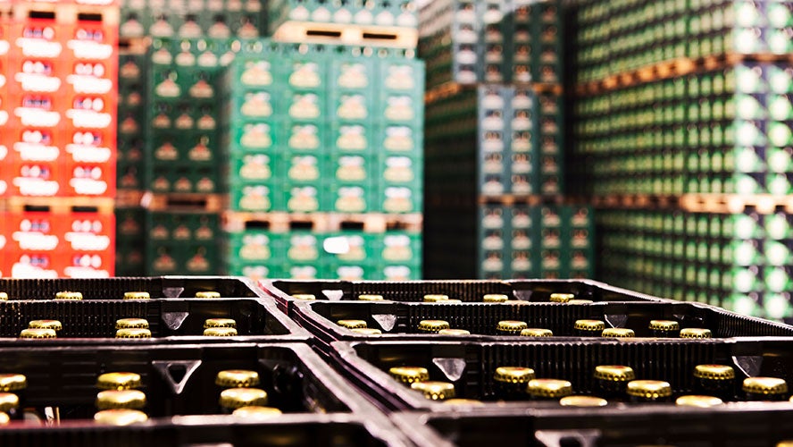 Stacks of crates filled with bottled drinks, gold bottle caps gleaming in the foreground, highlight the food and beverage supply chain in a warehouse-like setting, with colorful bottles blurred in the background.