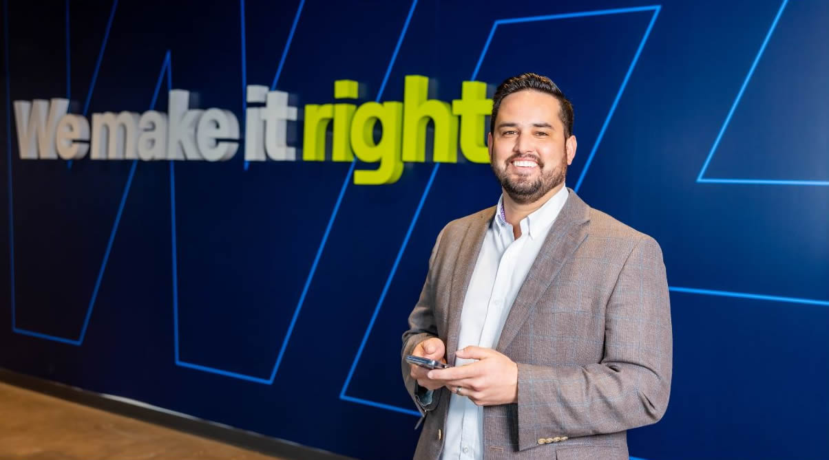 A man in a gray blazer smiles and holds a smartphone, standing in front of a blue wall with the slogan We make it right in large letters—reflecting the commitment of a leading 3PL company.