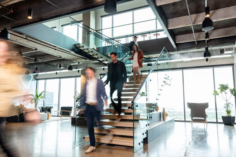 Four people in business attire move energetically around a modern 3PL company office, with three descending a glass staircase and one walking by, surrounded by large windows and indoor plants.