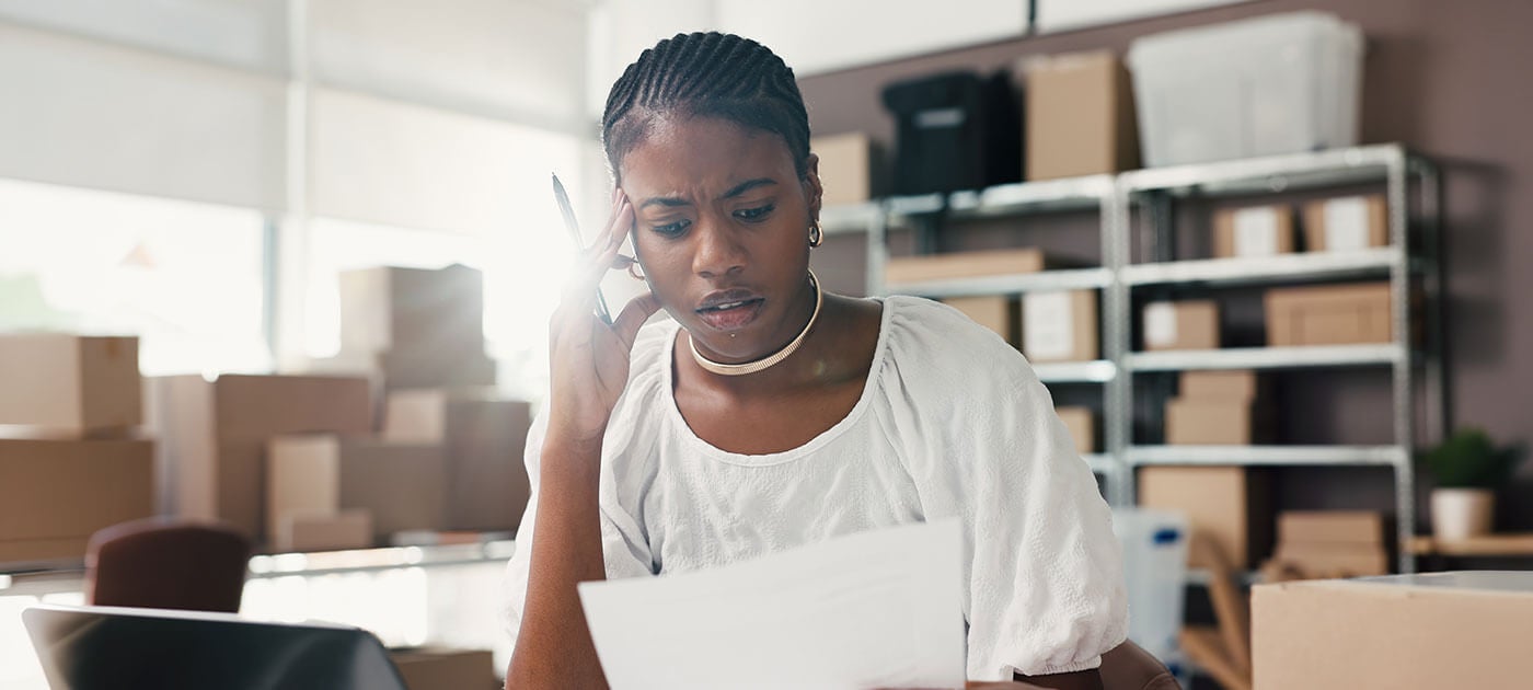A woman sits at a desk surrounded by boxes, looking concerned while reading a document and holding a pen to her temple, possibly stressed about work or finances.