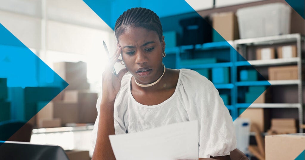 A woman holding a pen looks concerned while reading a document in a workspace with boxes and shelves in the background.