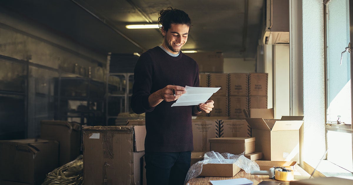 A smiling person stands in a warehouse or storage room, holding papers amid cardboard boxes, tape, and packing materials on a table near a window—perhaps conducting a freight bill audit to ensure accuracy and efficiency in the shipping process.