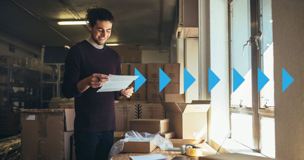 A person smiles while holding a piece of paper in a room with cardboard boxes. Blue arrows point right, leading toward a window, suggesting a shipping or order fulfillment process—potentially following a freight bill audit to ensure accuracy before dispatch.