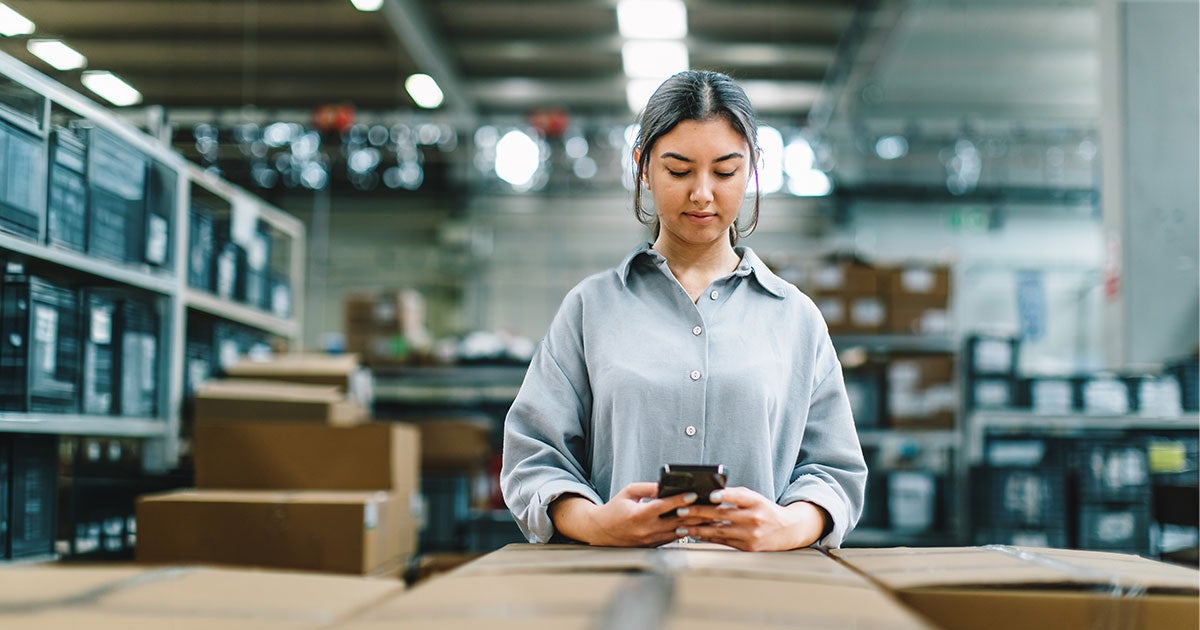 A woman in a warehouse stands among boxes, looking down at a smartphone—perhaps reviewing key performance indicators for supply chain management. Shelves filled with more boxes are visible in the background.