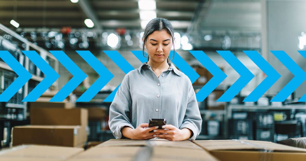 A person stands in a warehouse, looking at a phone, surrounded by cardboard boxes, with blue arrows overlaid across the image—highlighting the importance of tracking supply chain KPIs for efficient operations.