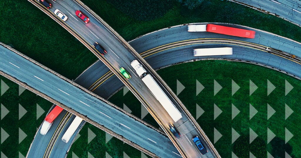 Aerial view of intersecting highways with cars and trucks driving on curved roads over green grass, showing motion blur on some vehicles, including transport trucks used to ship large items across long distances.