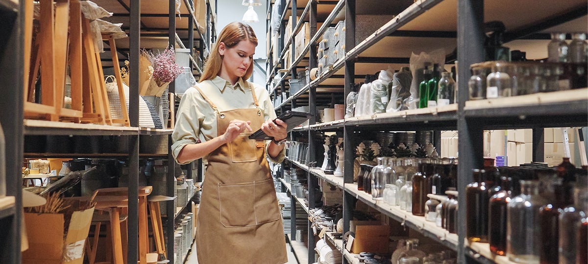 A person wearing an apron stands in a storage room surrounded by shelves of jars, bottles, and household items, using a tablet to check inventory and review tariff refund information.