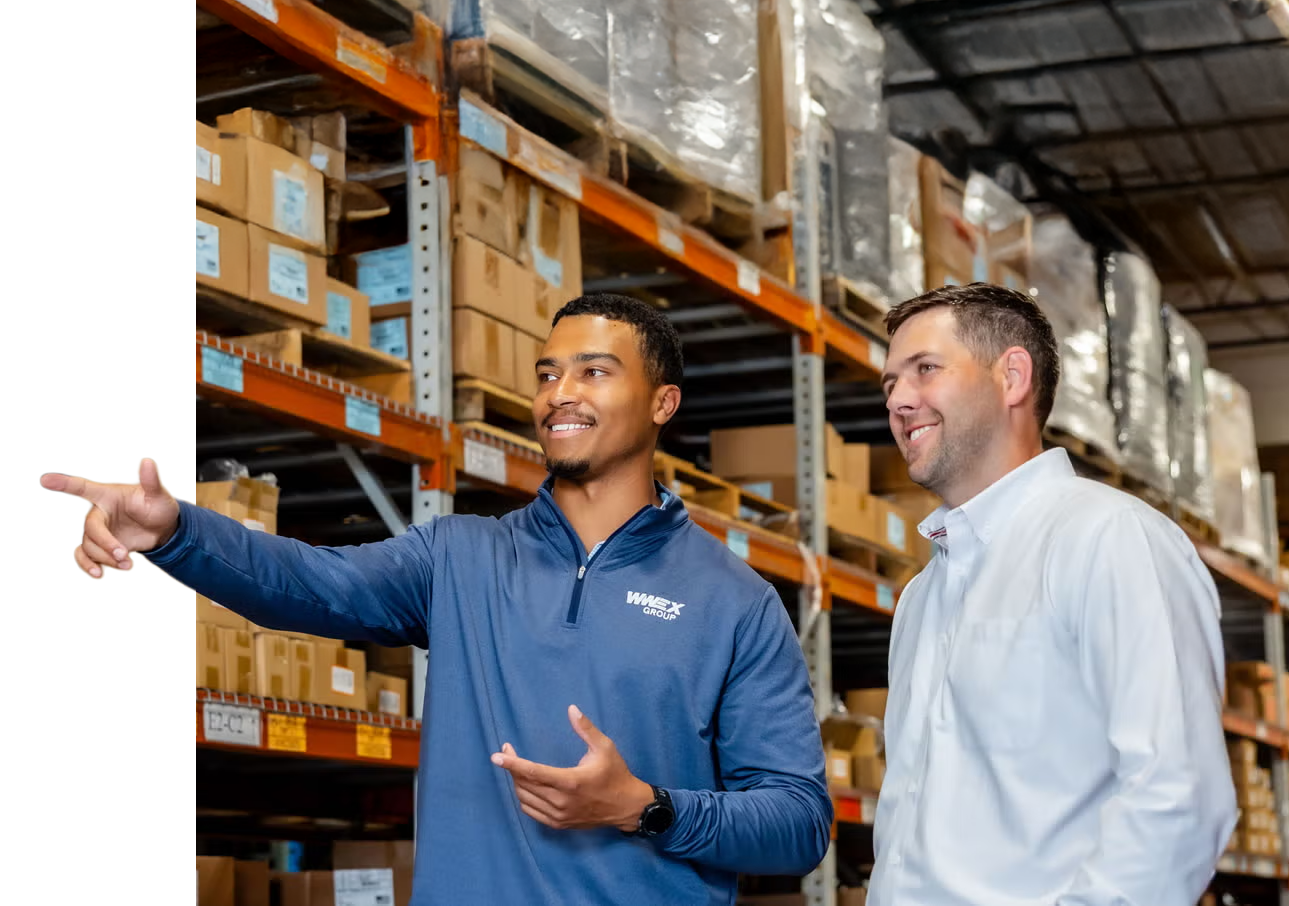 Two men stand in a warehouse full of shelves and boxes. One man, wearing a blue shirt, points and smiles while talking to the other man, who wears a white shirt. Both appear engaged in conversation.