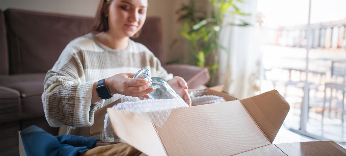 A person carefully packs a glass jar into a cardboard box with bubble wrap—demonstrating best practices for shipping fragile items—while sitting on a couch in a sunlit living room with plants in the background.