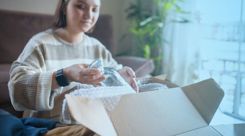 A person wearing a striped sweater and smartwatch carefully unpacks a glass item—clearly marked as shipping fragile items—from a cardboard box filled with packing peanuts, sitting on the floor in a bright room with a couch and plants in the background.