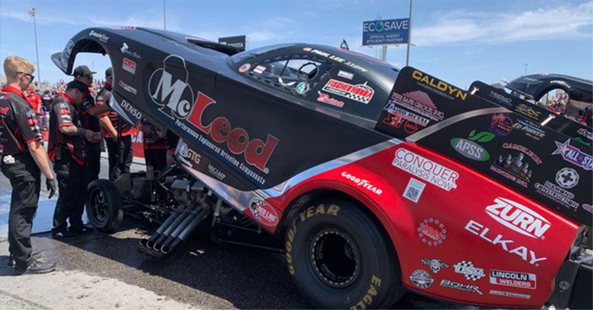 Crew members work on a black and red drag race car with its body lifted, exposing the engine, at a racetrack. Showcasing the precision often seen in manufacturing logistics, the car is covered in sponsor logos and parked on asphalt under a sunny sky.