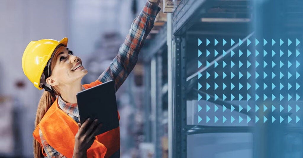 A smiling worker wearing a yellow hard hat and orange vest uses a tablet to check nmfc codes while reaching for an item on a high shelf in a warehouse. Blue triangular graphics appear on the right side of the image.