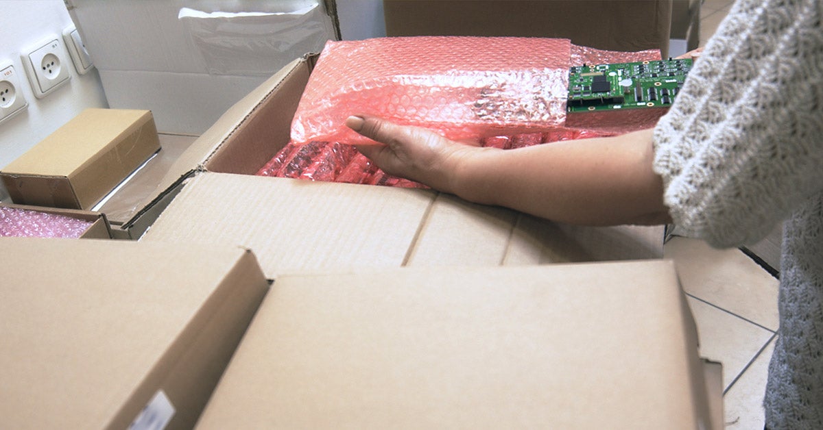 A person’s hand places a green circuit board wrapped in pink bubble wrap into a cardboard box for shipping electronics.