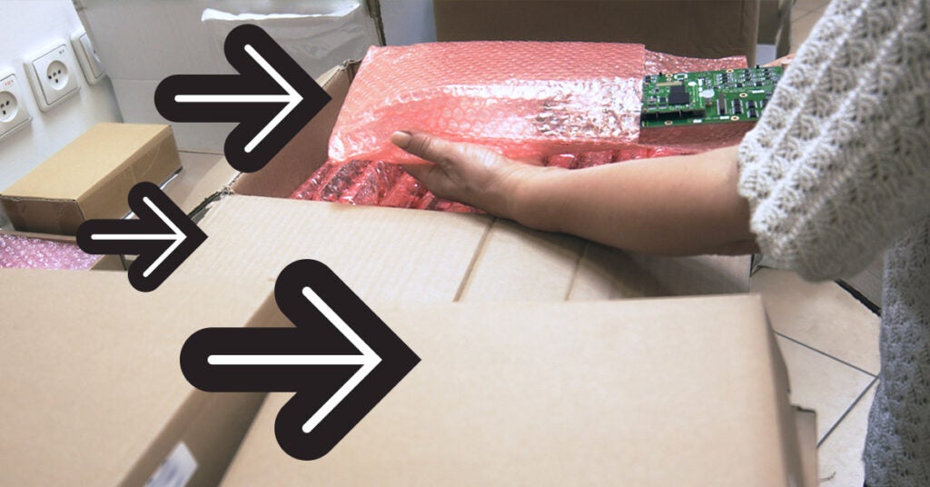A hand places a green circuit board wrapped in pink bubble wrap into a cardboard shipping box for electronics.
