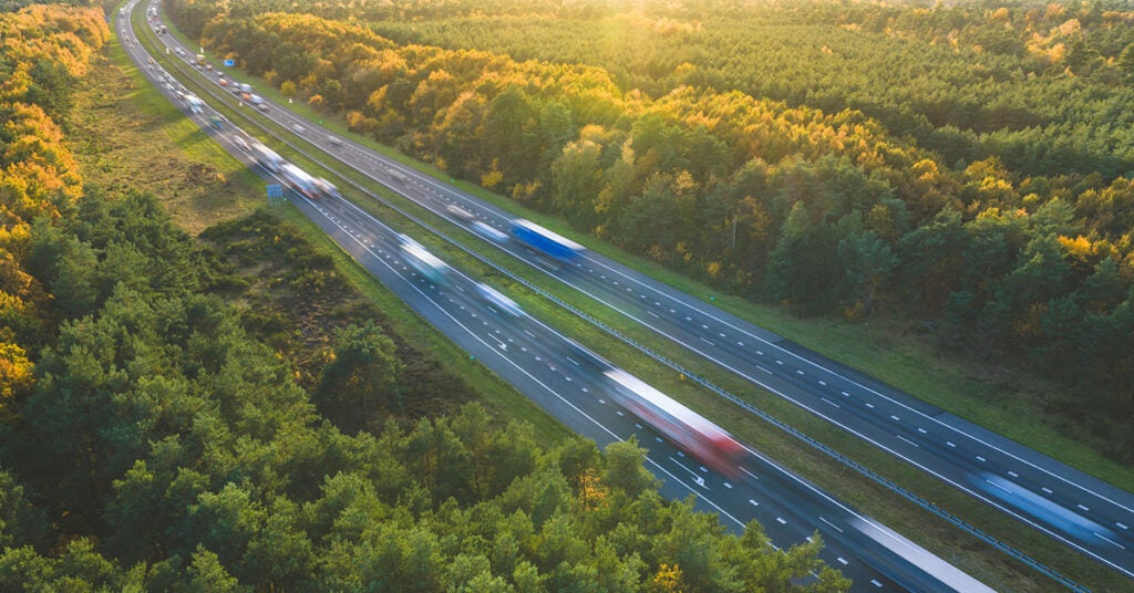 An aerial view of a highway transporting shipping electronics through an autumn forest, with motion-blurred trucks driving in both directions.
