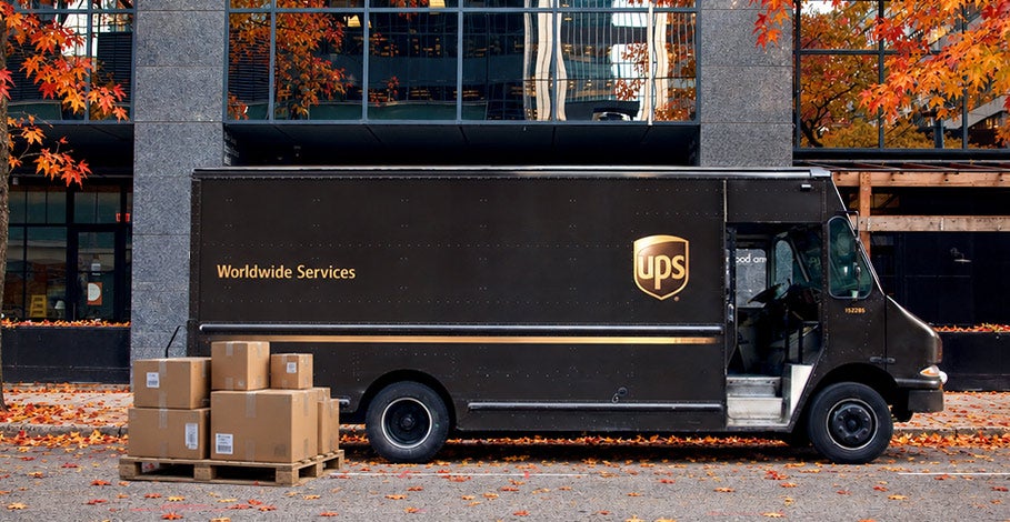 A brown UPS delivery truck parked on a street covered in fallen autumn leaves during freight peak season, with a wooden pallet holding cardboard boxes placed on the ground nearby.