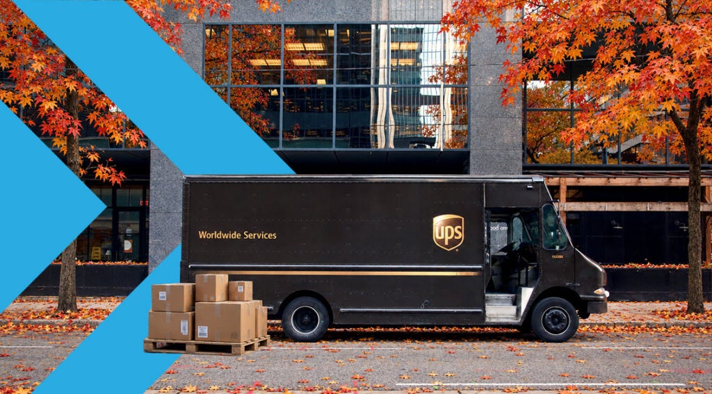 A brown UPS delivery truck parked on a street with fall foliage during the freight peak season, with a pallet of cardboard boxes on the ground nearby.